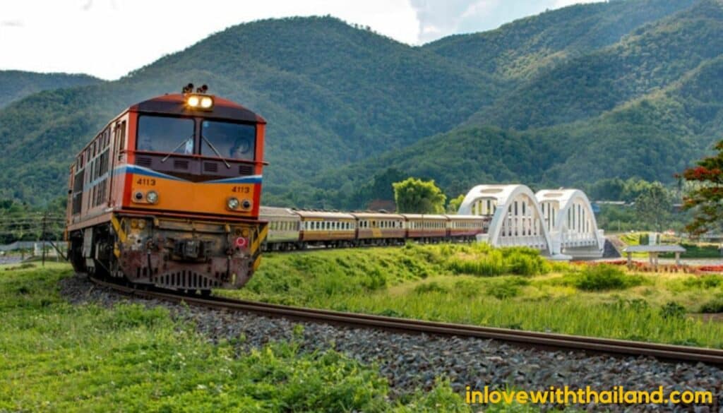 Train going from Bangkok to Chiang Mai  through a green scenery. It has a white bridge in the background and mountains.